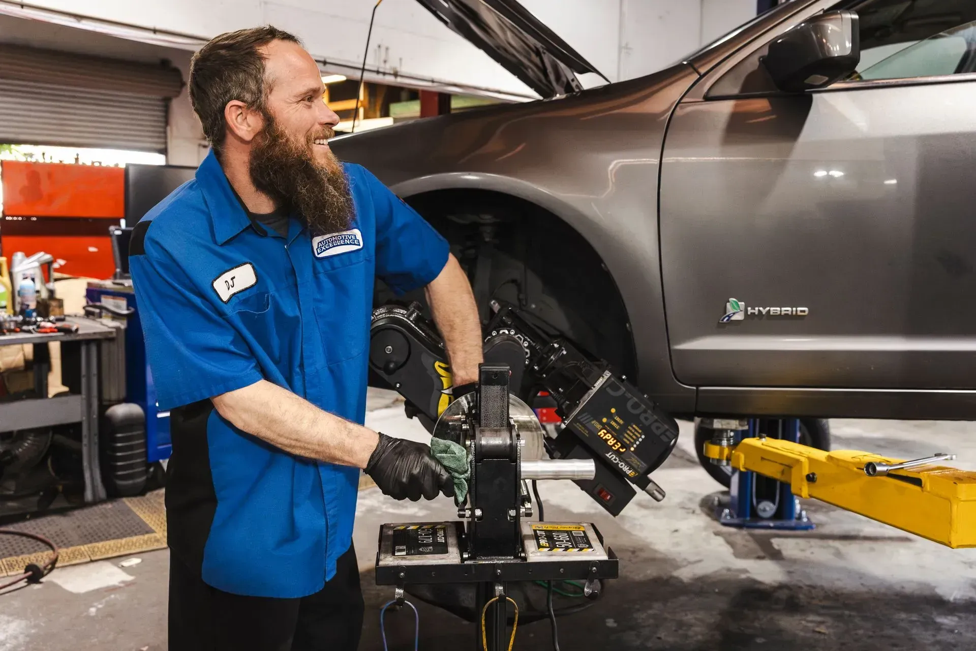 Mechanic working on car's suspension with a tool in a shop. He wears a blue shirt and smiles. | Automotive Excellence