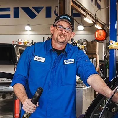Mechanic in blue uniform, smiling, holding hose, standing in auto repair shop. | Automotive Excellence