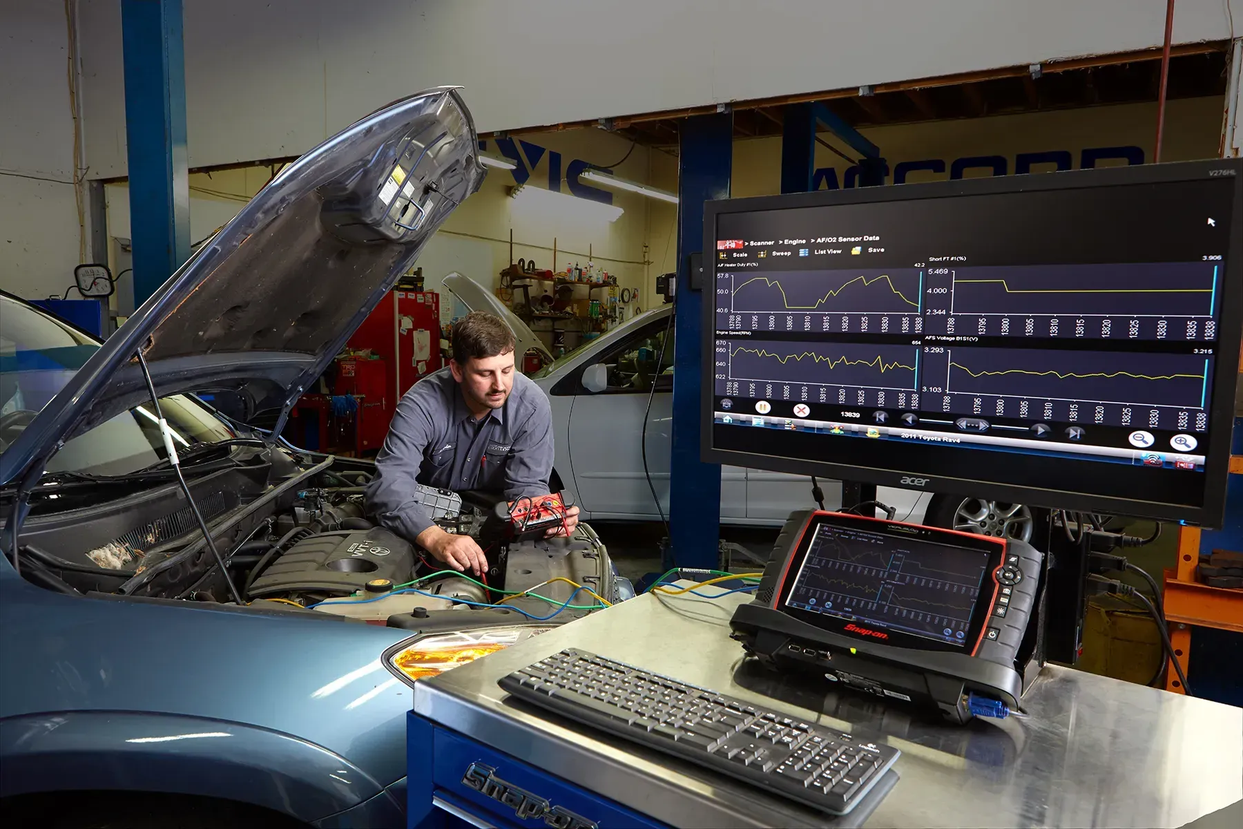 Mechanic working on car engine, monitoring diagnostics on large screen. Shop setting, blue car, tools. | Automotive Excellence