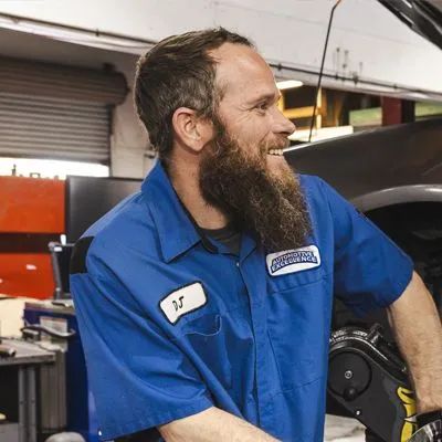 Mechanic in blue uniform smiles, leaning near a car in a garage. | Automotive Excellence
