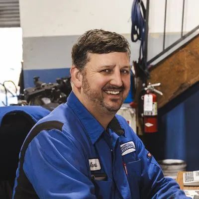 Man in blue mechanic's uniform smiling, in a garage setting with tools and equipment. | Automotive Excellence