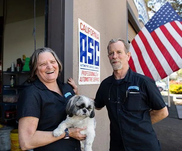 Woman holding dog, man in front of auto shop, California Automotive Service sign, American flag. | Automotive Excellence
