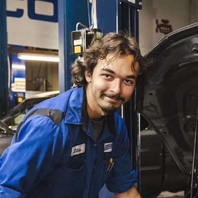 Mechanic in blue coveralls smiles, leaning against a car with open hood in a garage. | Automotive Excellence