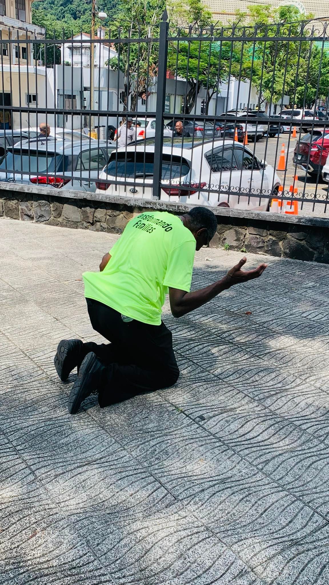 Man kneels on ground, wearing neon shirt, appears to be praying outdoors near a fence and parked cars.