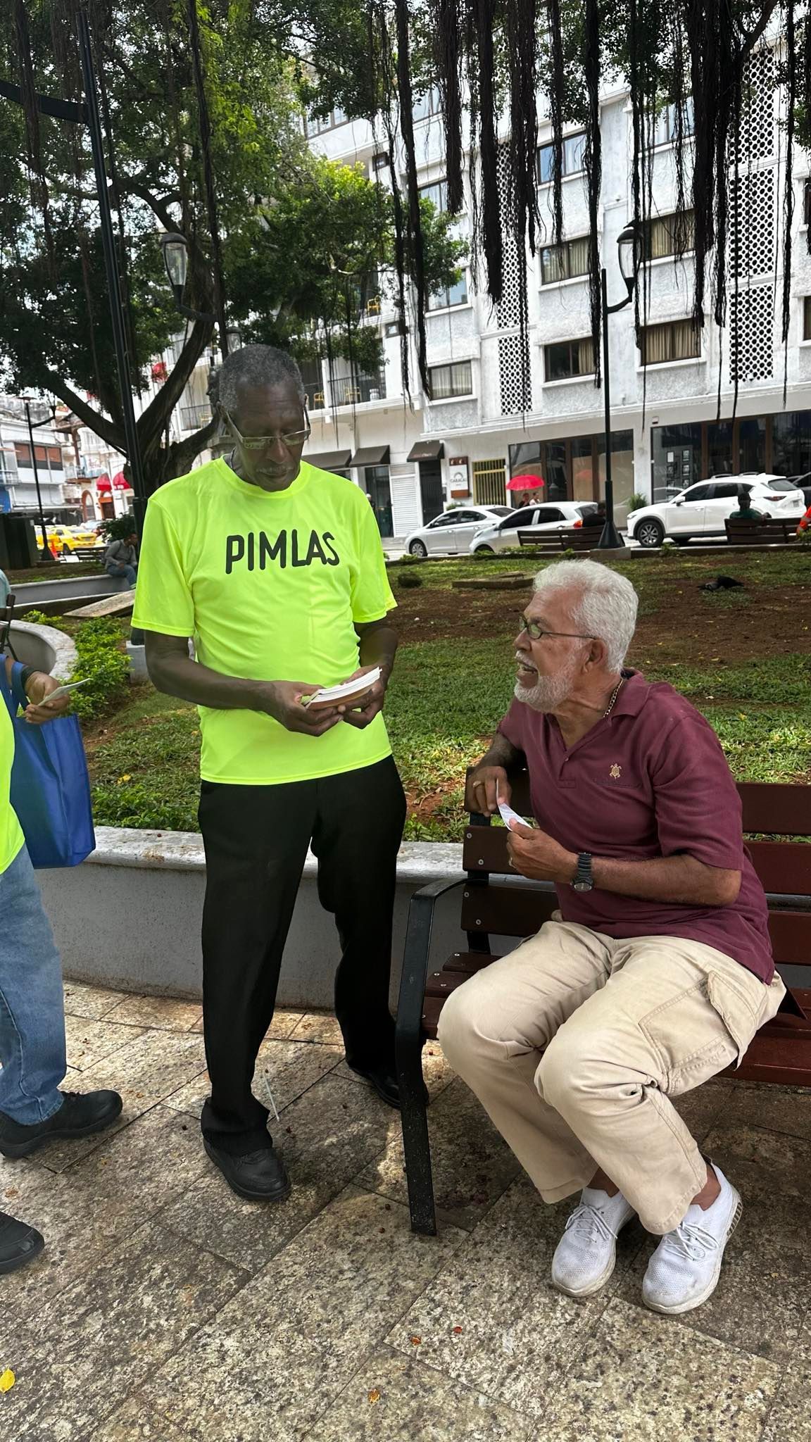 Two men in a park; one standing, one seated, both holding paper items. Buildings and trees in the background.
