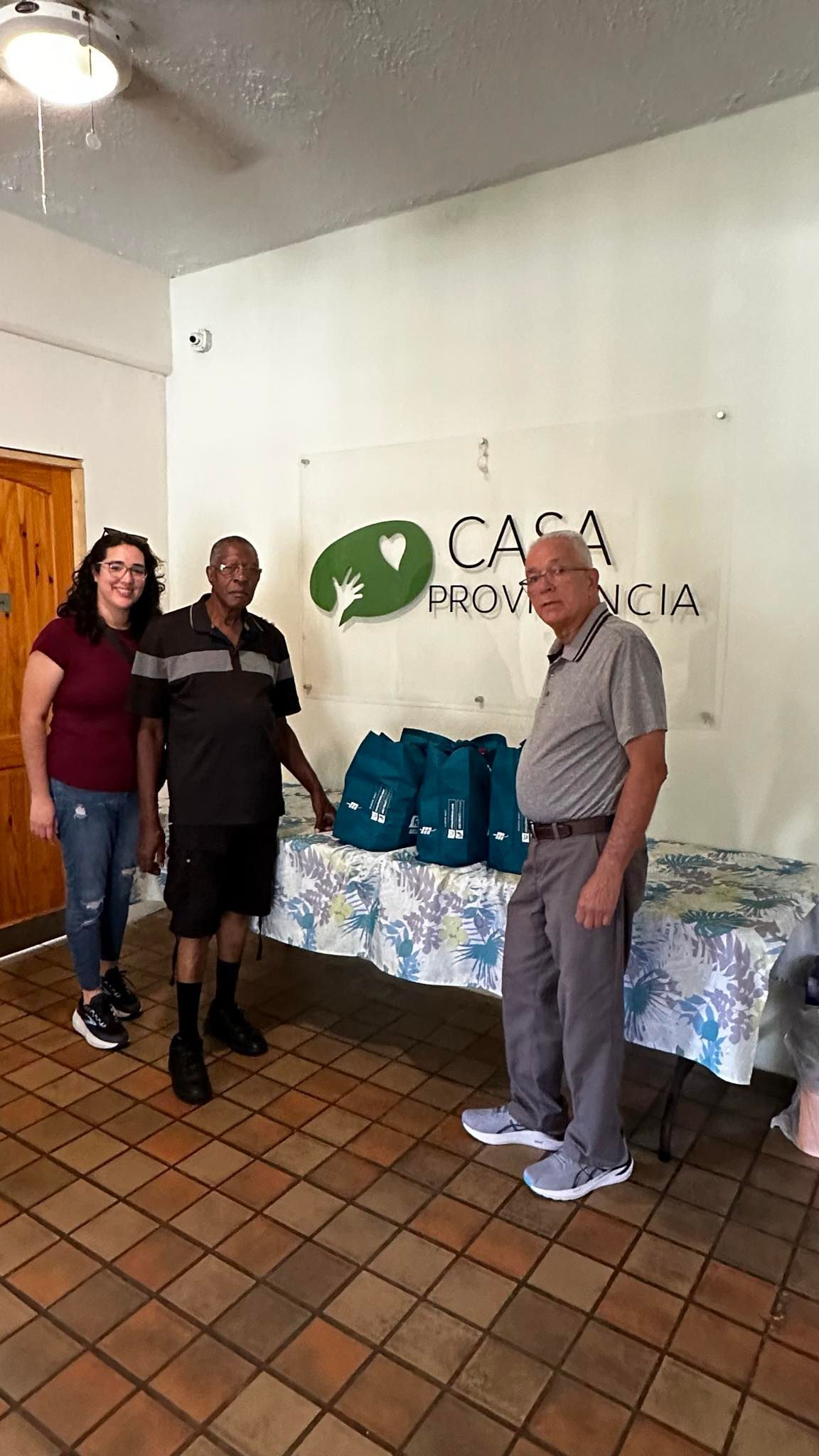 Three people stand by a table with gifts, near a sign that says 