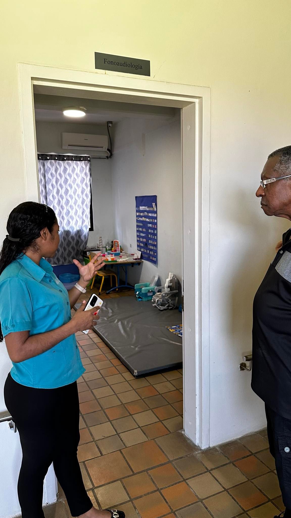 Woman in teal shirt speaks to man by a doorway to a room with supplies and a sign.