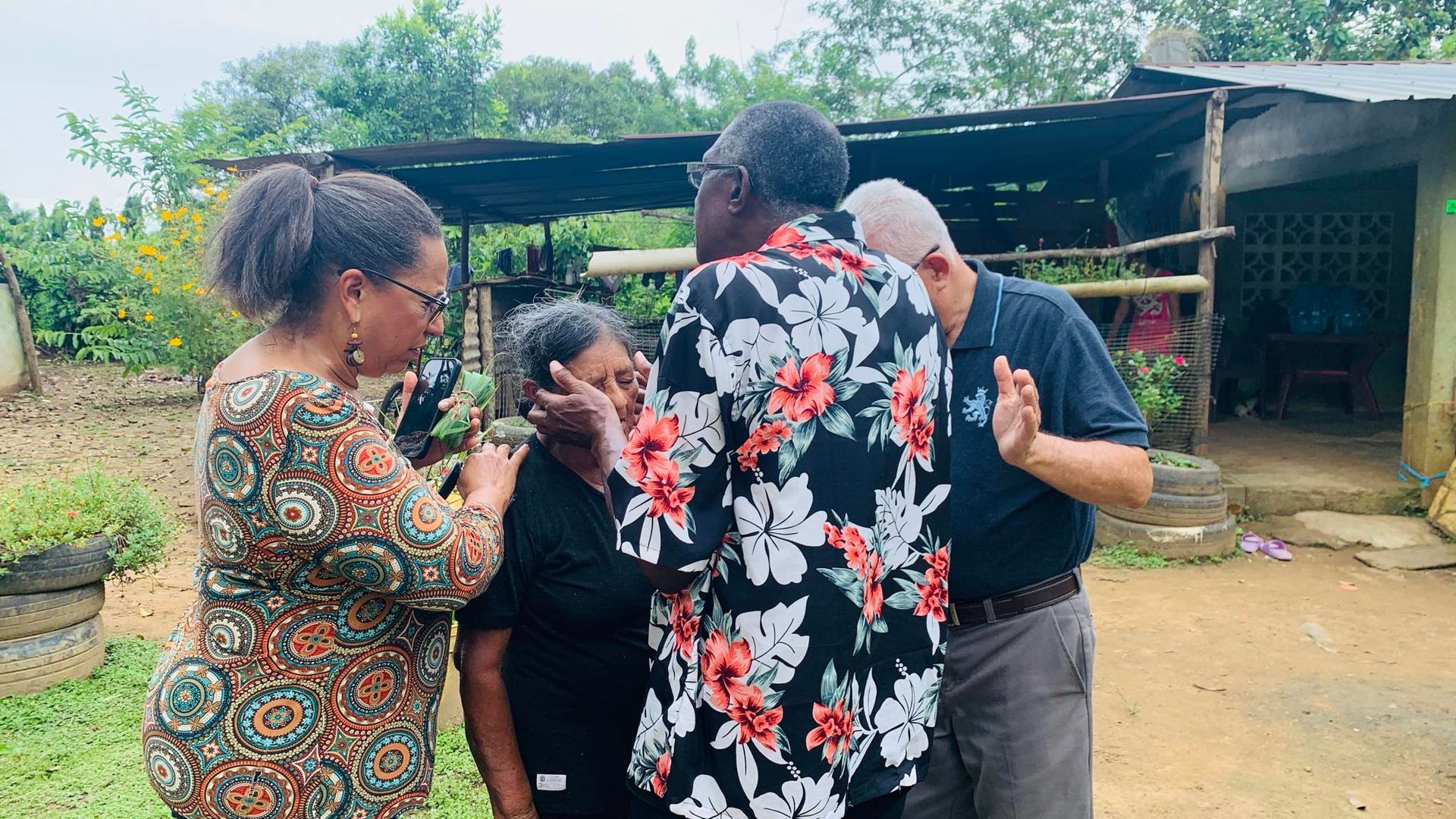 Four people in front of a house: woman being comforted; others praying, touching shoulders.