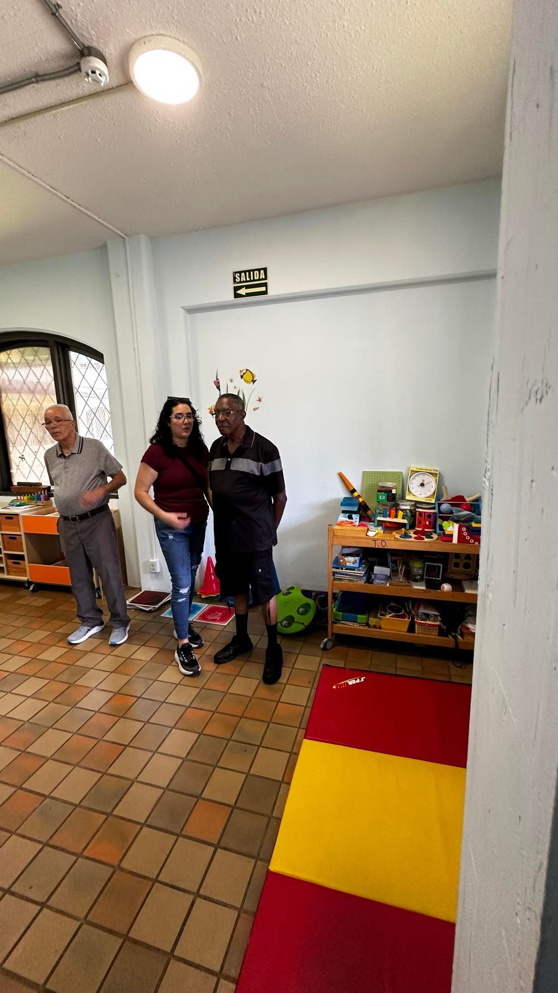Three people inside a room, two looking at each other. Red, yellow, and orange floor mat visible.