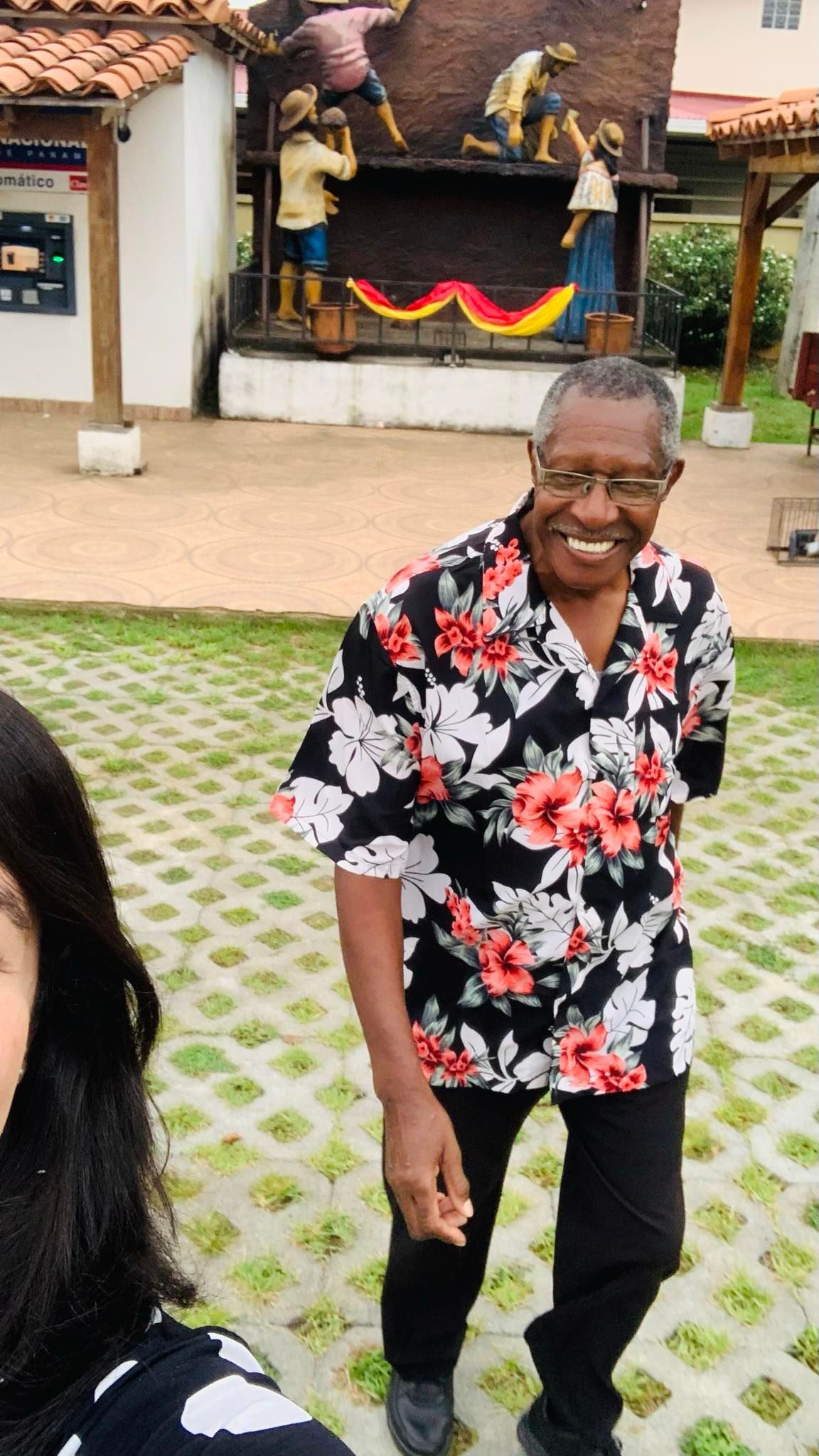 Smiling older Black man in floral shirt and black pants poses outdoors with a woman; background has statues.