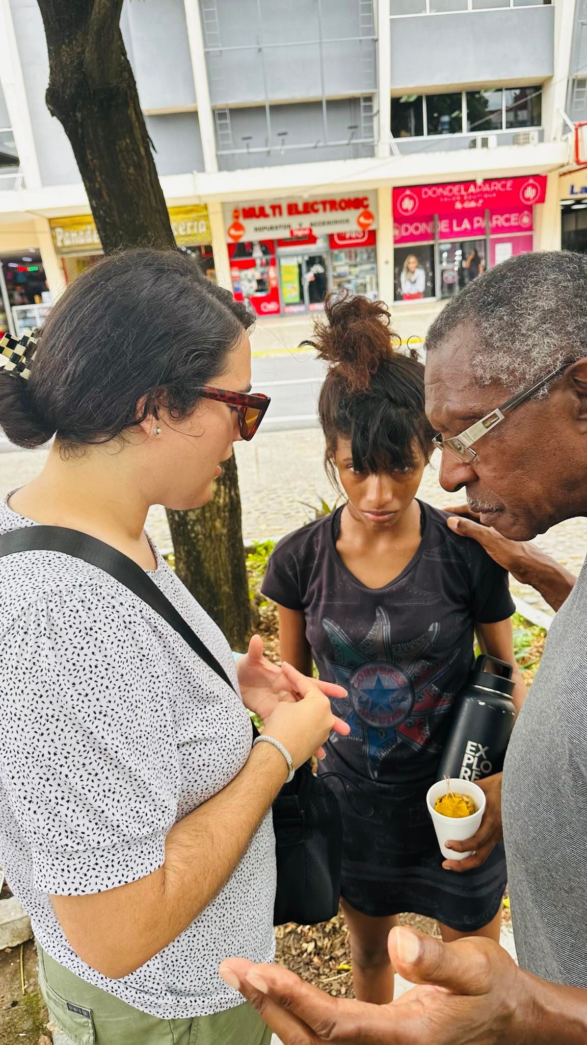 Three people standing together, two older adults talking to a younger person. City setting, daytime.