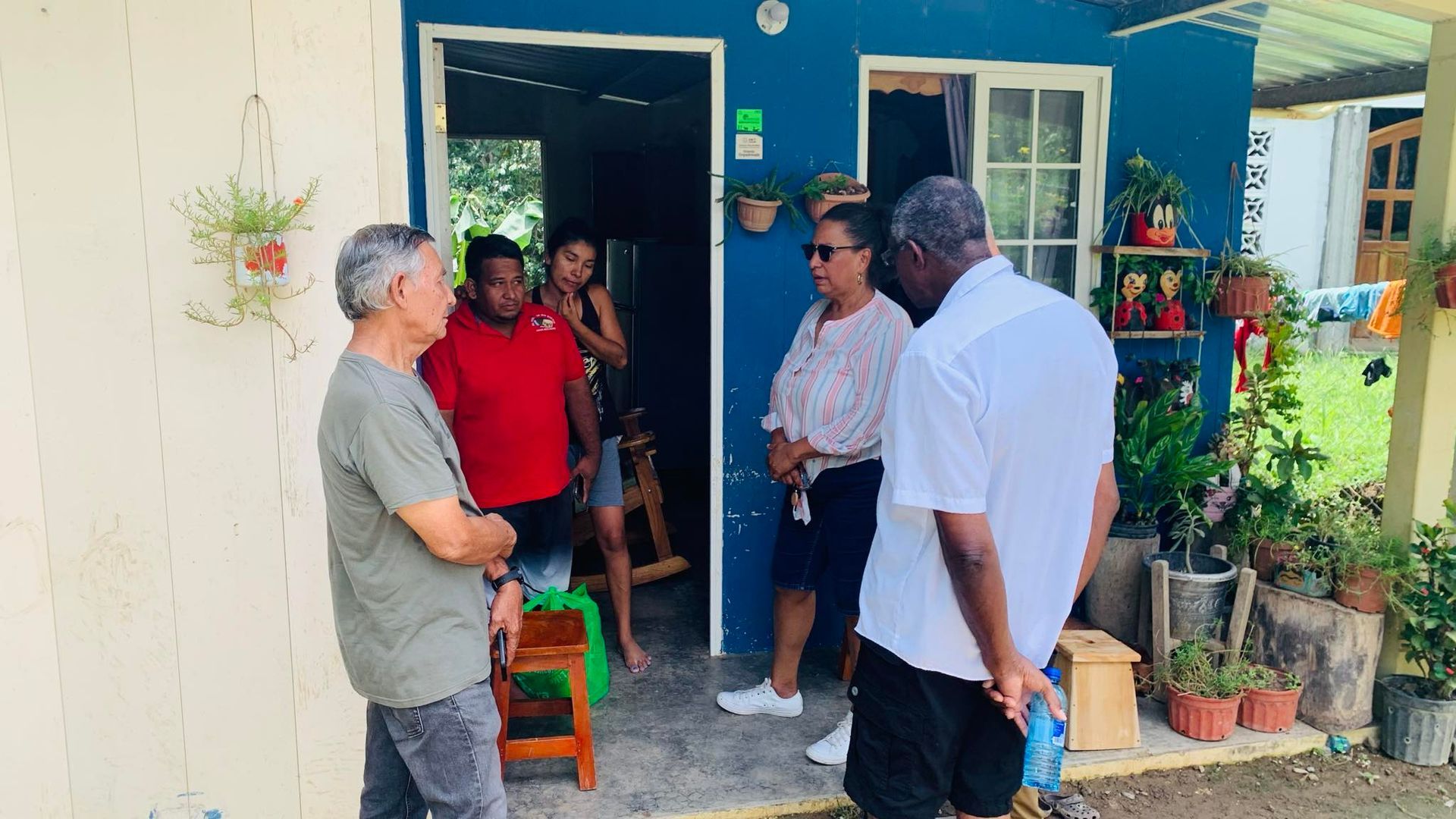 People gathered outside a blue house with open door. Several are talking; plants decorate the porch.