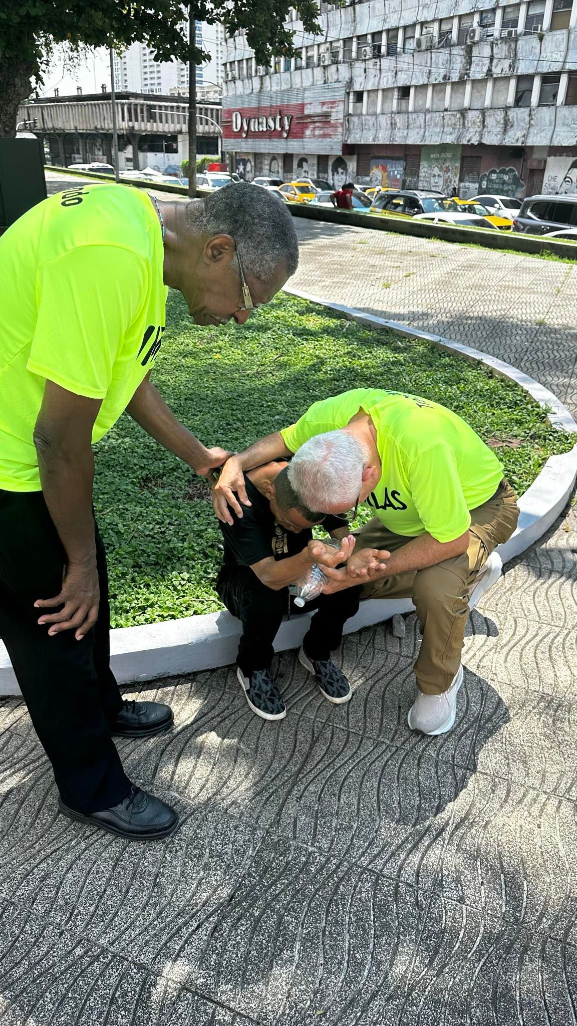 Three people in neon yellow shirts outdoors: one helping two others, one kneeling, one leaning.