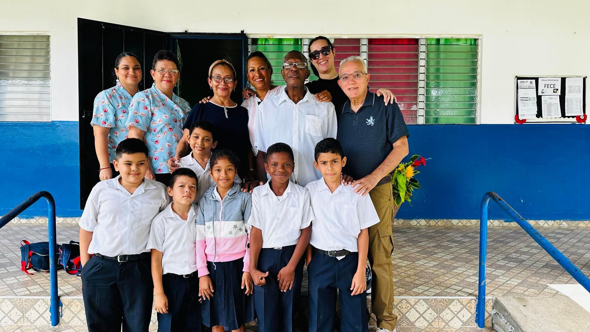 Group photo: diverse adults with children in front of a school building; smiling, posing; bright colors.