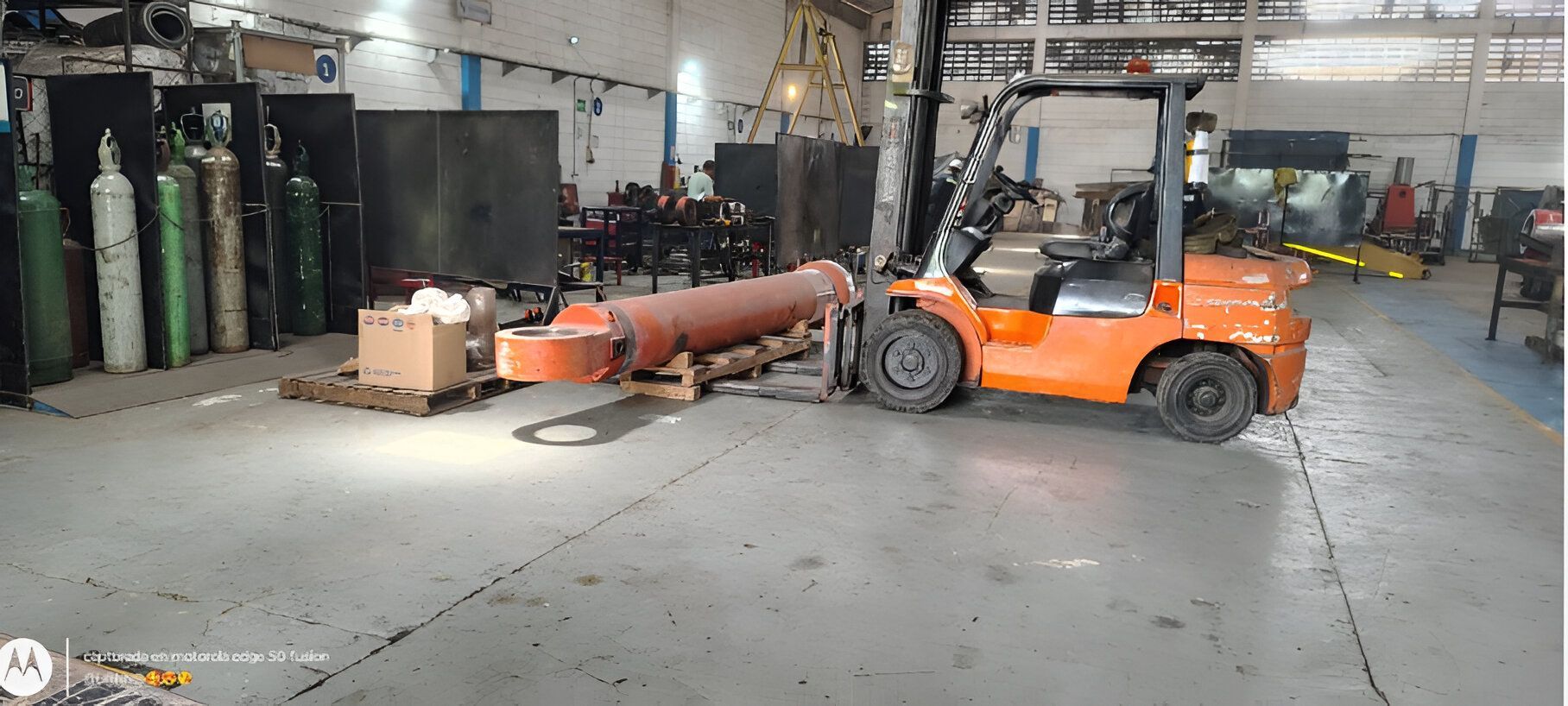 Una carretilla elevadora naranja transporta un gran objeto cilíndrico en un almacén.