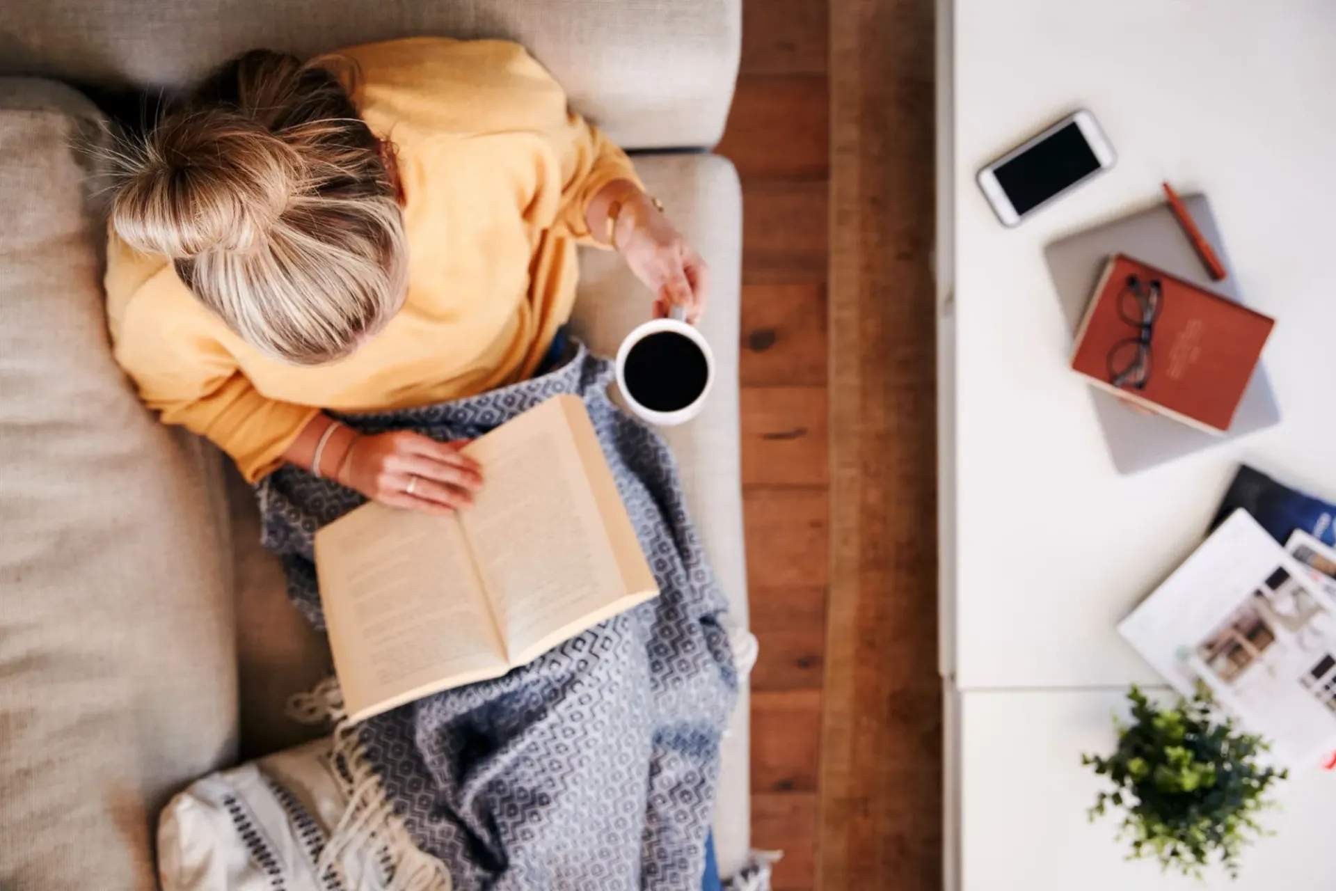 Person reading a book on a couch beside a coffee table with a tablet, notebook, and plant