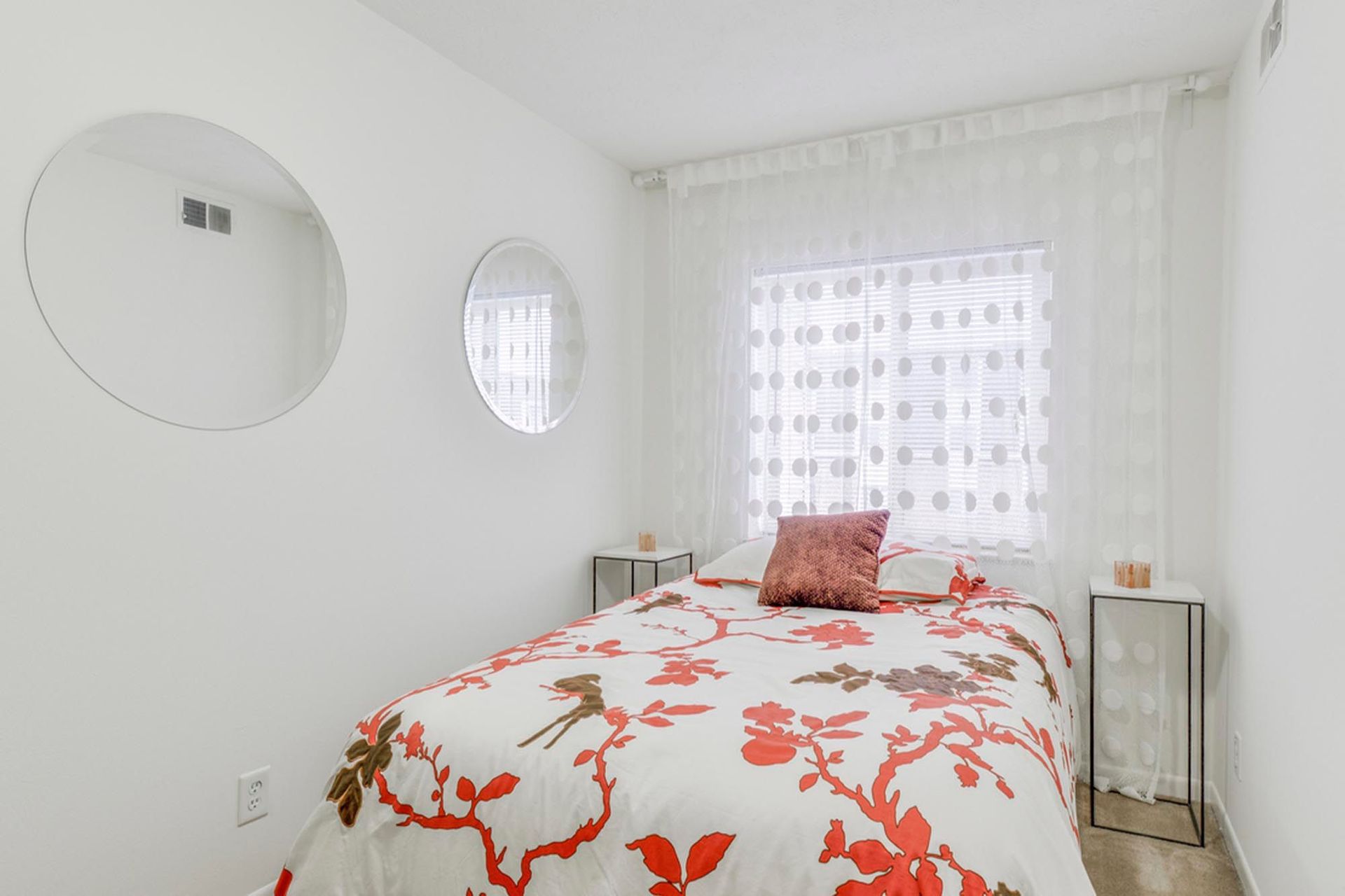 Small white bedroom with a single bed, floral duvet, round mirrors, and sheer polka-dot curtains at the Reserves at Arlington in Columbus, OH.