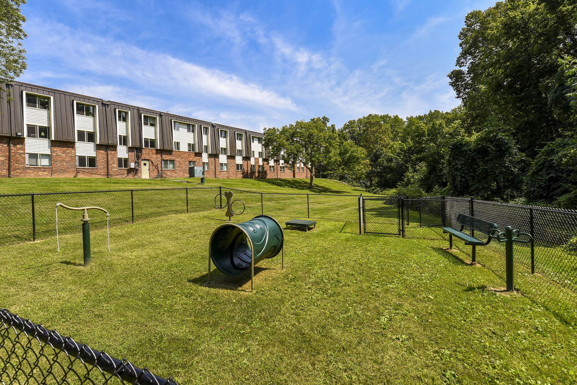 Fenced grassy community playground with a tunnel slide, a bench, and an apartment building in the background at the Reserves at Arlington in Columbus, OH.