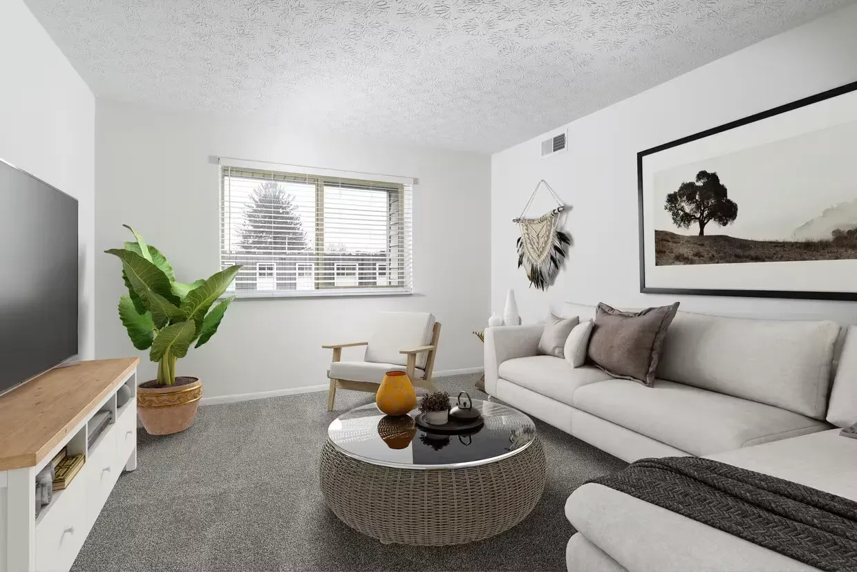 Bright apartment living room with gray sectional sofa, round glass-top wicker coffee table, plant, and wall art at the Reserves at Arlington in Columbus, OH.