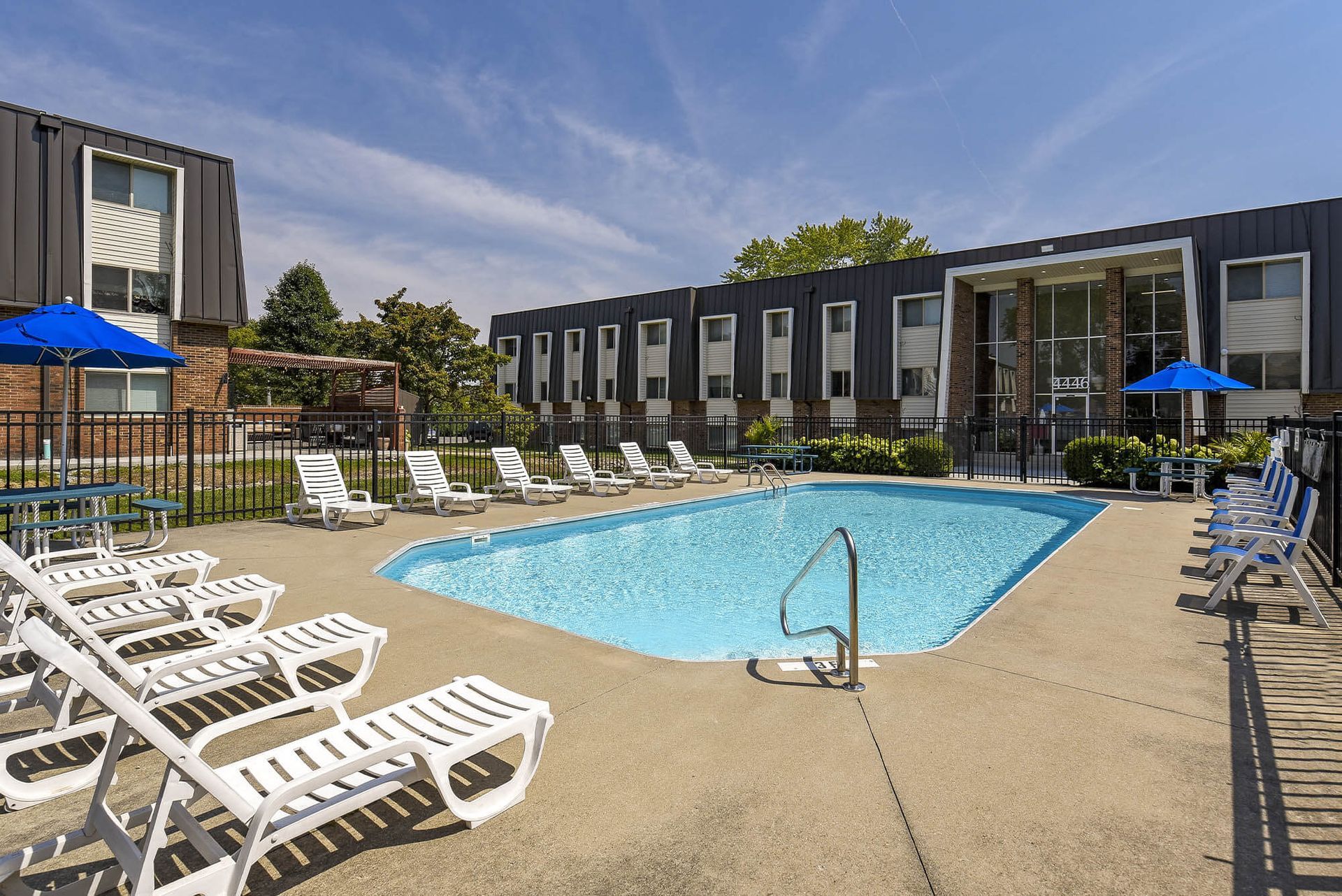 Outdoor pool area with white lounge chairs, blue umbrellas, and a gated fence beside modern apartment buildings at the Reserves at Arlington in Columbus, OH.
