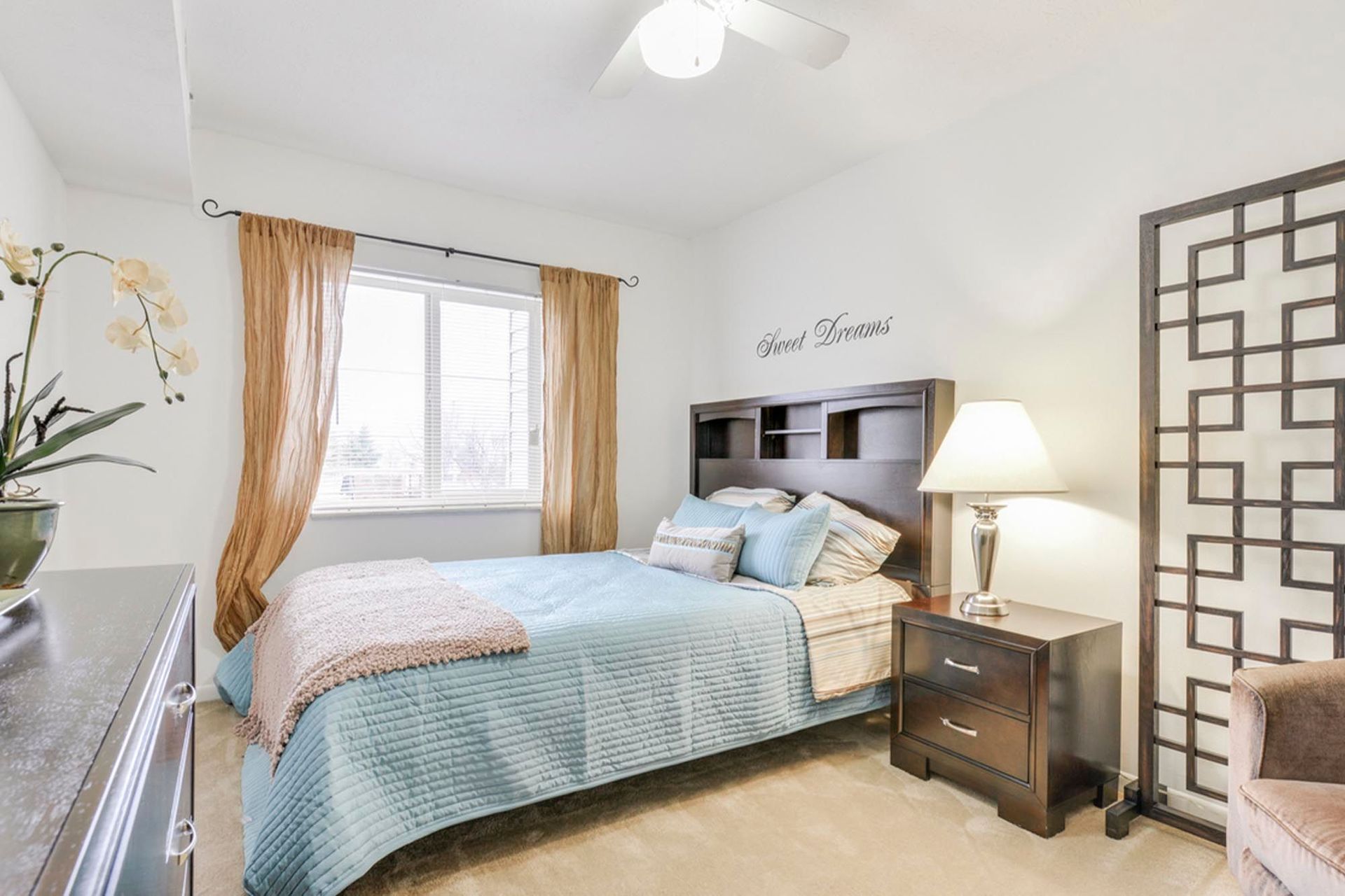 Cozy apartment bedroom with a queen bed, dark wood headboard, nightstand, lamp, and window with tan curtains at the Reserves at Arlington in Columbus, OH.