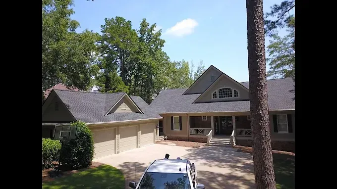 House with a dark roof and tan exterior, three-car garage, driveway, and trees under a sunny sky.