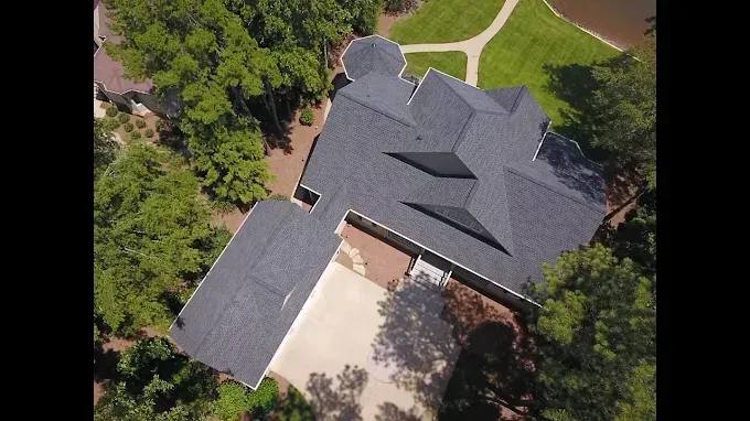 Aerial view of a large gray roofed house surrounded by trees, with a light colored driveway and a pond visible.