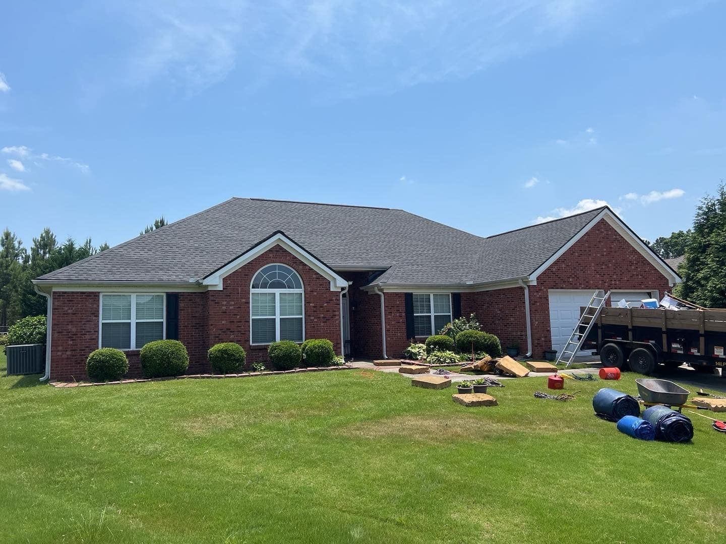 Brick house with a dark roof under a blue sky, surrounded by green grass and bushes; roofing materials in yard.