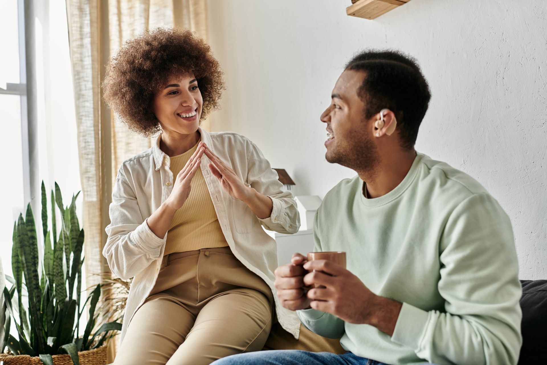 Woman gesturing excitedly, talking to a man holding a mug. They sit in a sunny room with a plant.