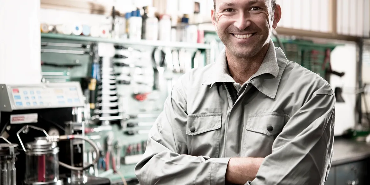 Mechanic with crossed arms smiles in workshop; tools and machinery in background. | Equality Automotive
