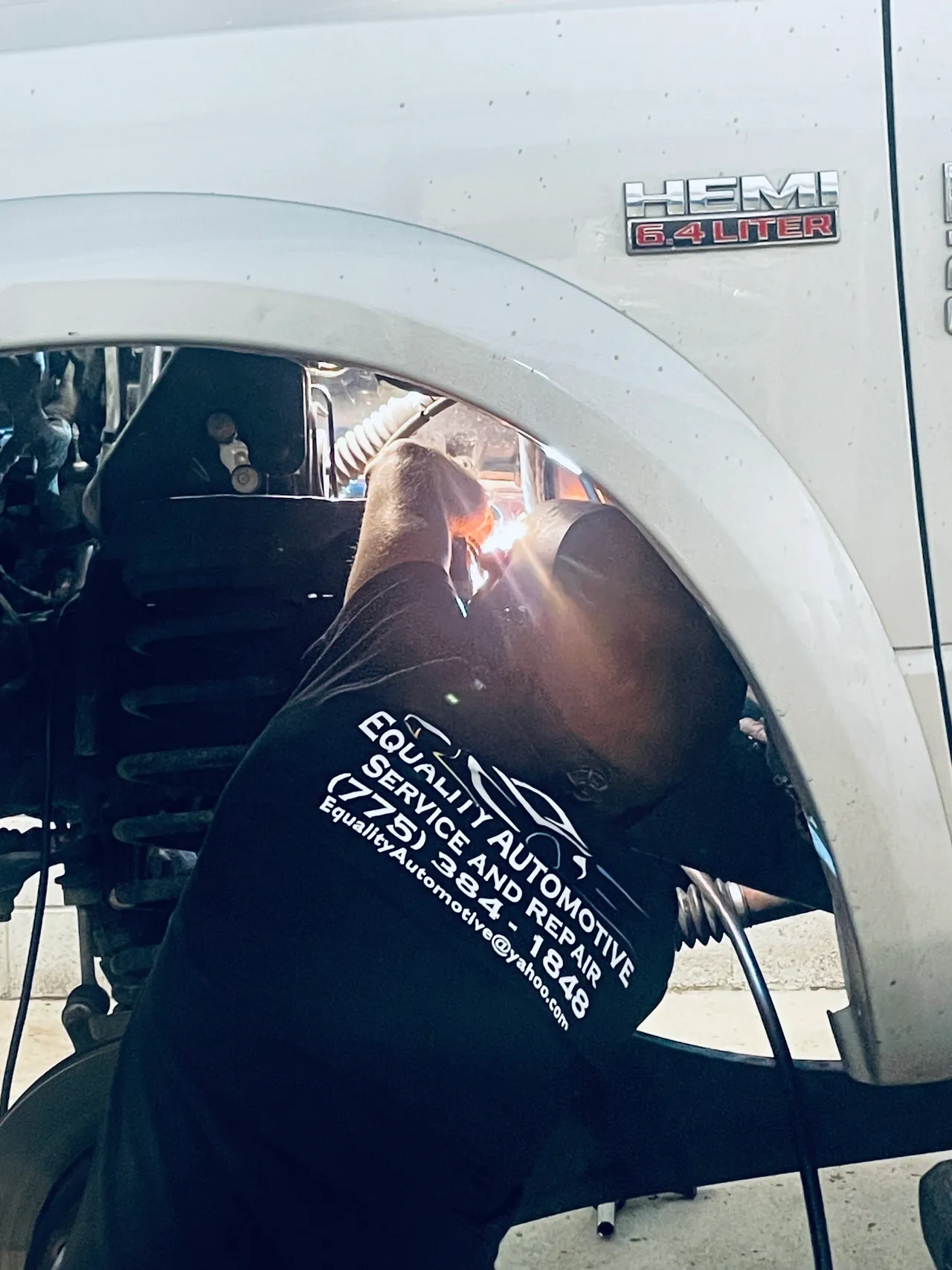 Mechanic welds a car part near the wheel, wearing a black shirt with business logo, outdoors. | Equality Automotive