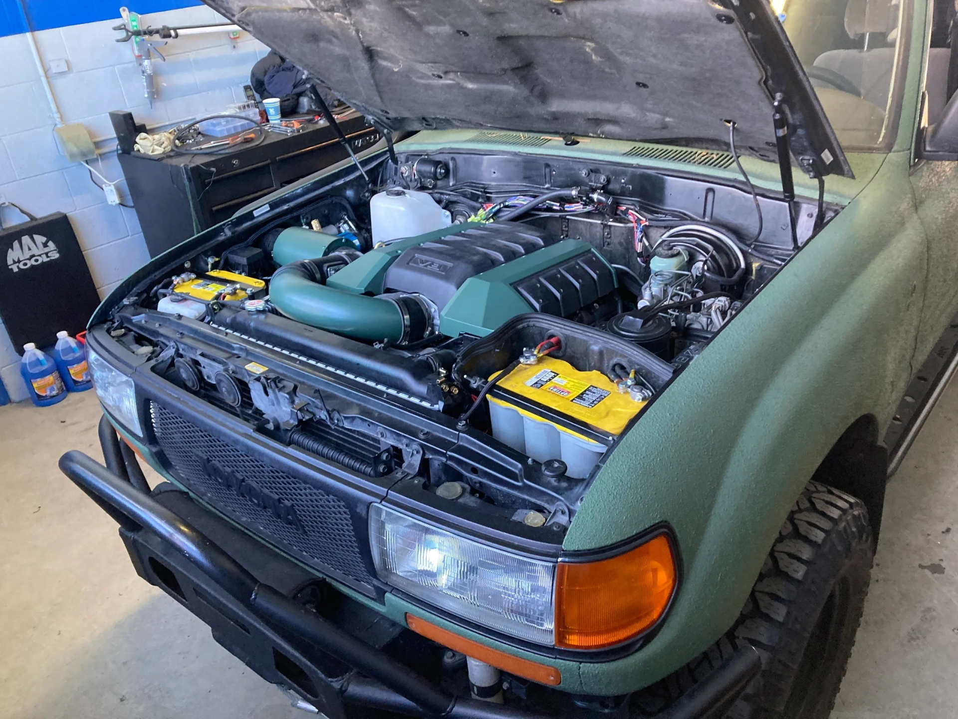 Green 80 series Land Cruiser with hood open, modified engine, black bumper, in a shop. | Equality Automotive