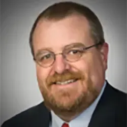 A man with a beard and glasses is wearing a suit and tie and smiling.