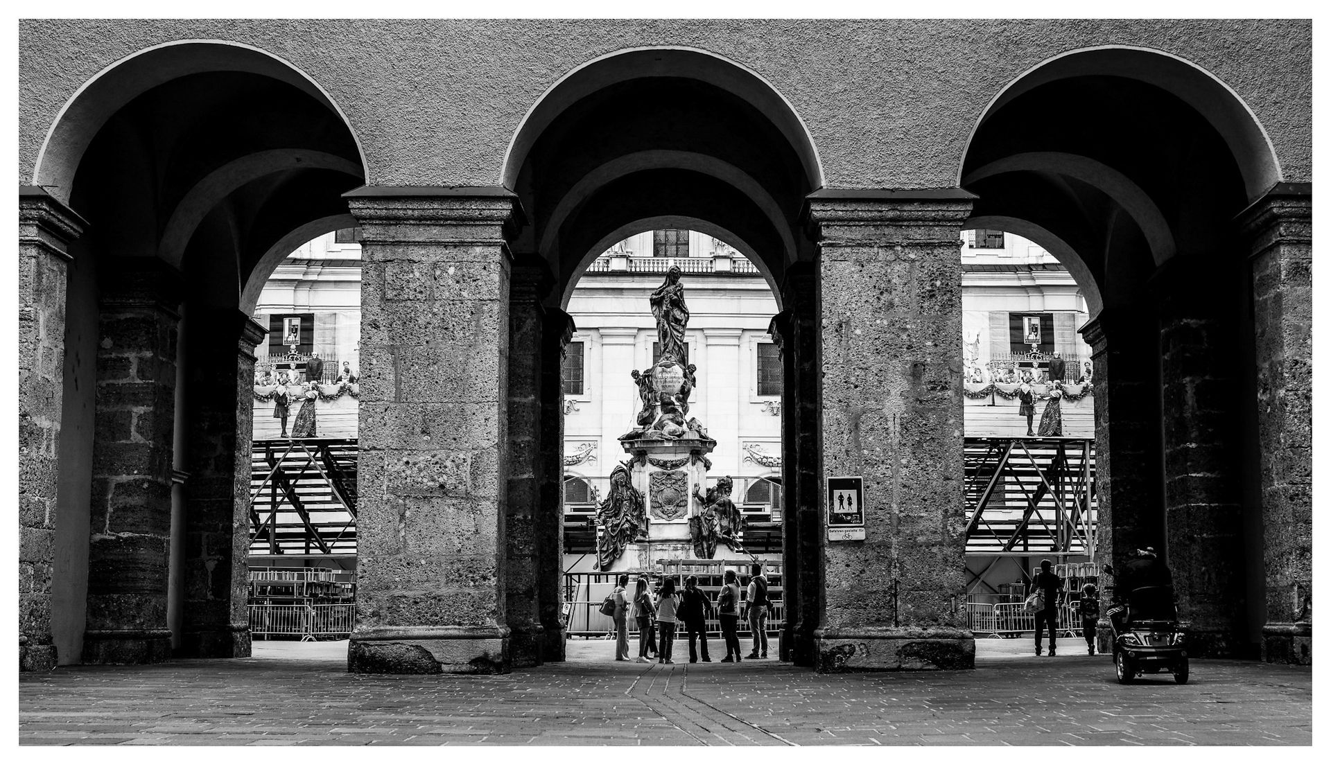 Three arched doorways frame a statue in a plaza. People gather near the statue, and scaffolding is visible.
