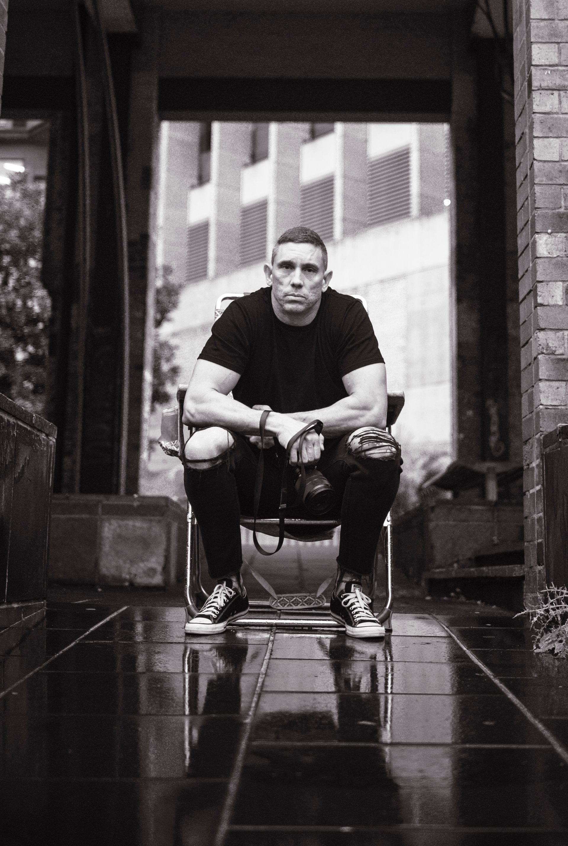 Man seated in a wheelchair, hands on lap, wearing black shirt and ripped jeans, outside. Black and white.