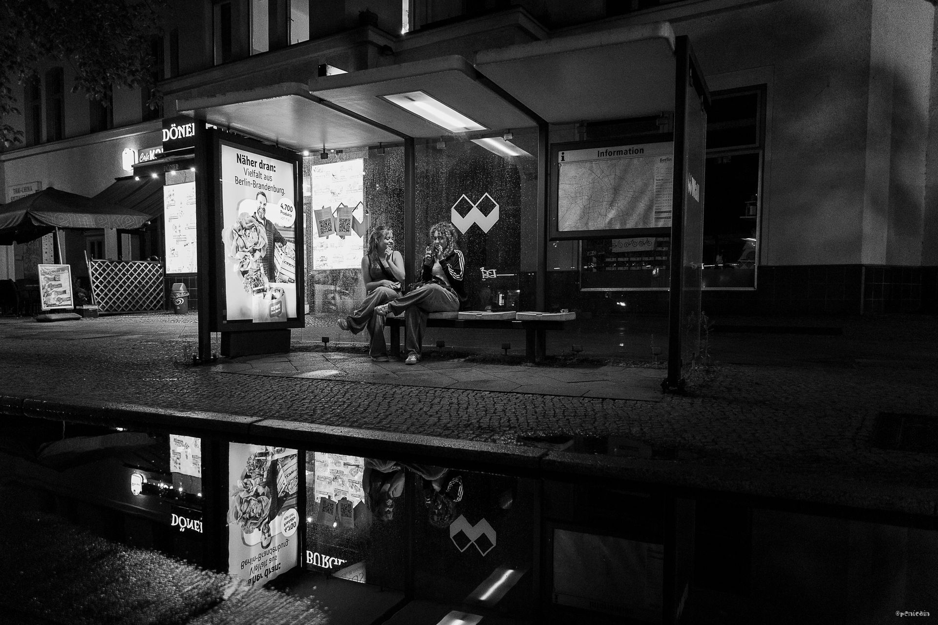 Bus stop at night, people sitting on a bench. Reflection in a puddle below.