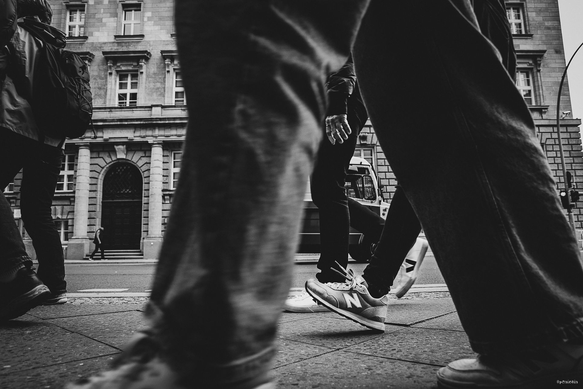 People walking on a city street, legs and feet in foreground, building in background. Black and white.