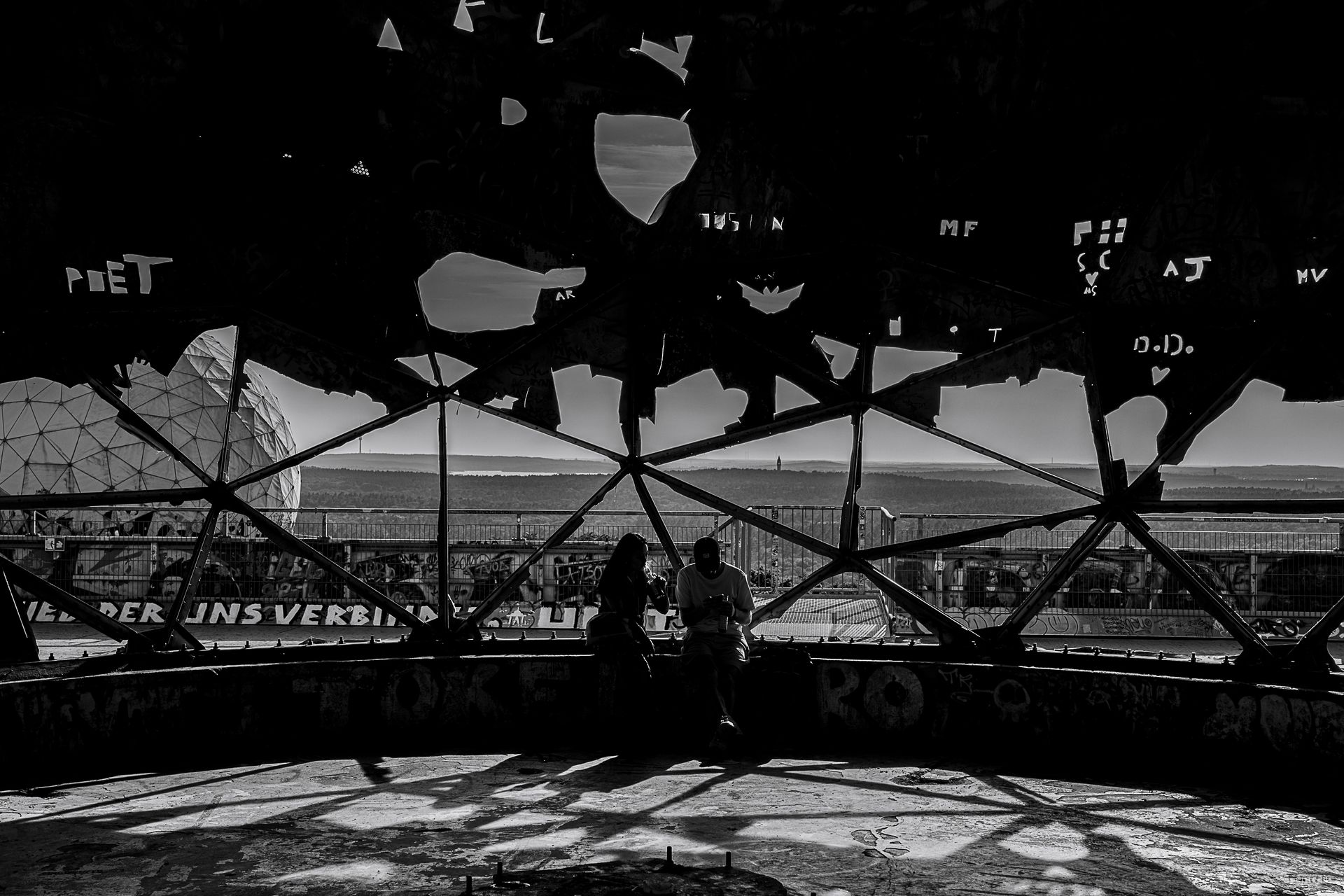 Inside a dome structure, two people silhouette against a landscape through geometric framework.