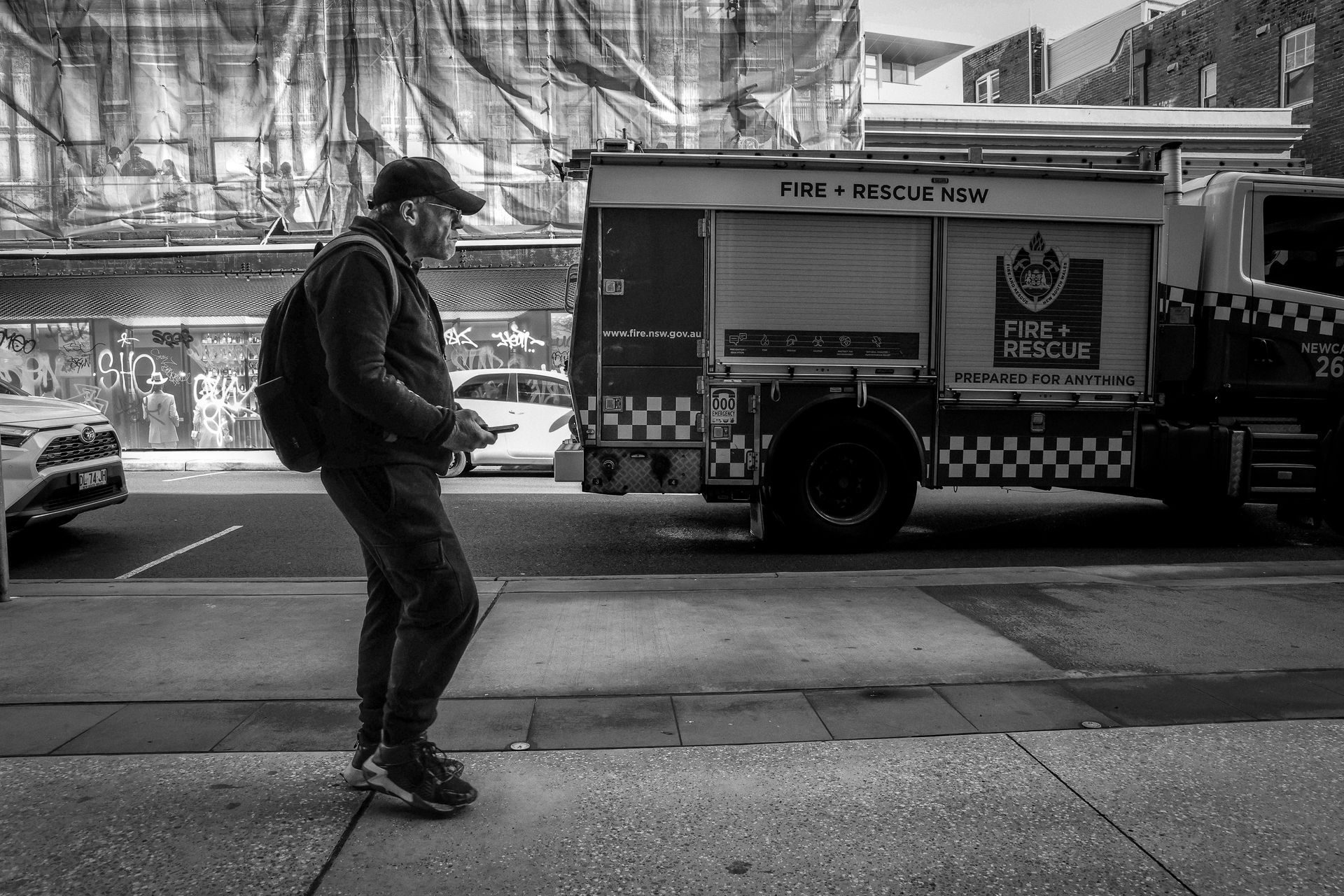 Person with backpack stands on sidewalk near a rescue truck, looking at phone. Street scene, black and white.