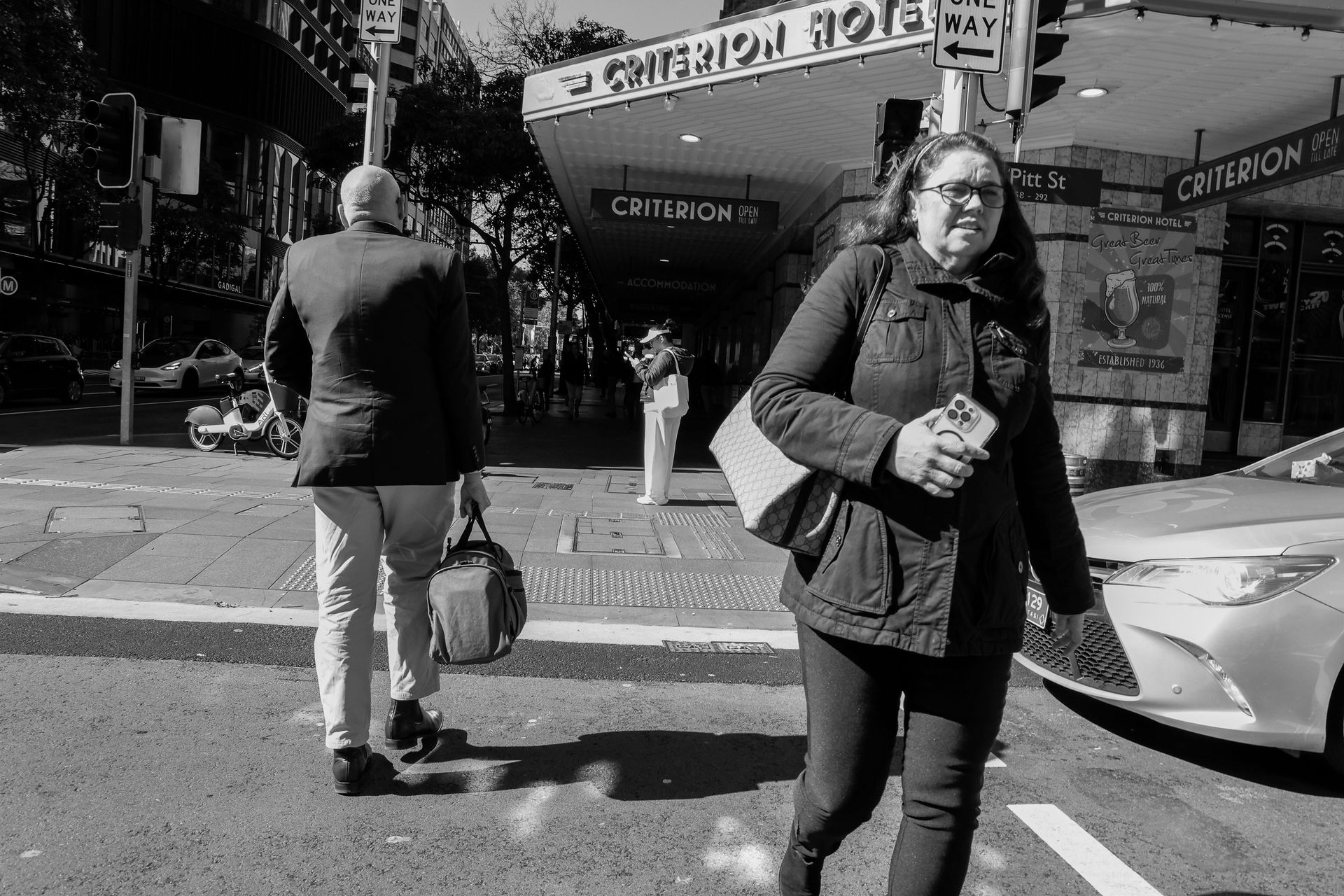 A pedestrian crosses a street, passing a man carrying a bag in front of a building with the words 