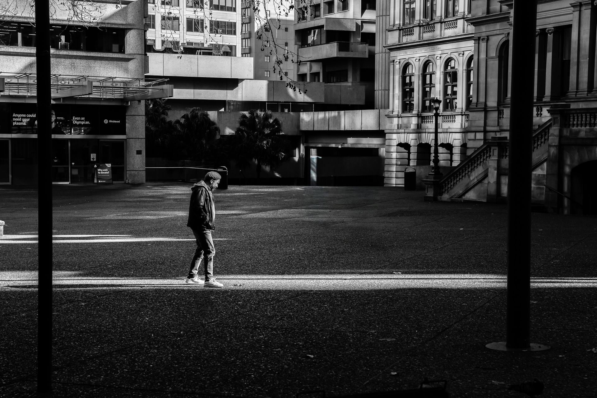Person walks across a paved plaza, framed by buildings and two vertical poles. Black and white photo.