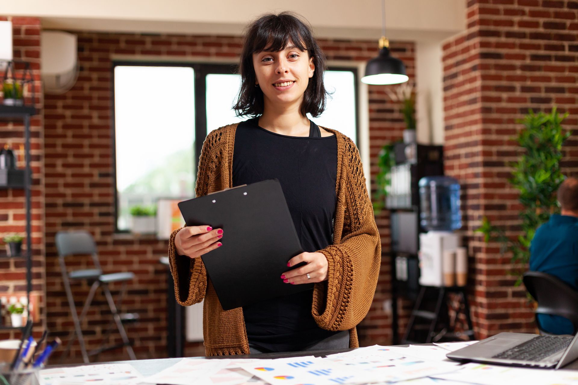 Woman holding a clipboard, smiling at the camera in an office setting with brick walls.