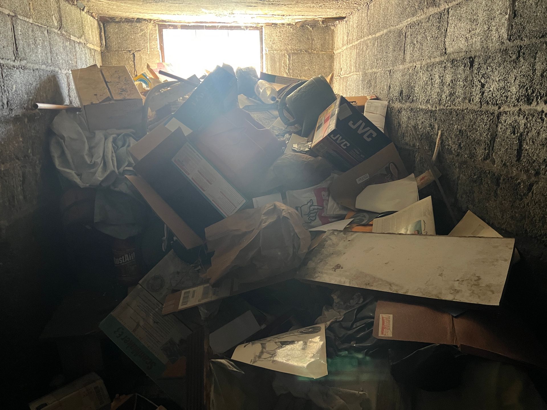 A dark, narrow storage space with cinder block walls, cluttered with cardboard boxes and debris, illuminated by a window.