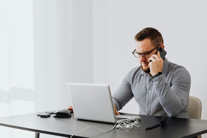 Man at a desk talking on a phone while working on a laptop in a bright office
