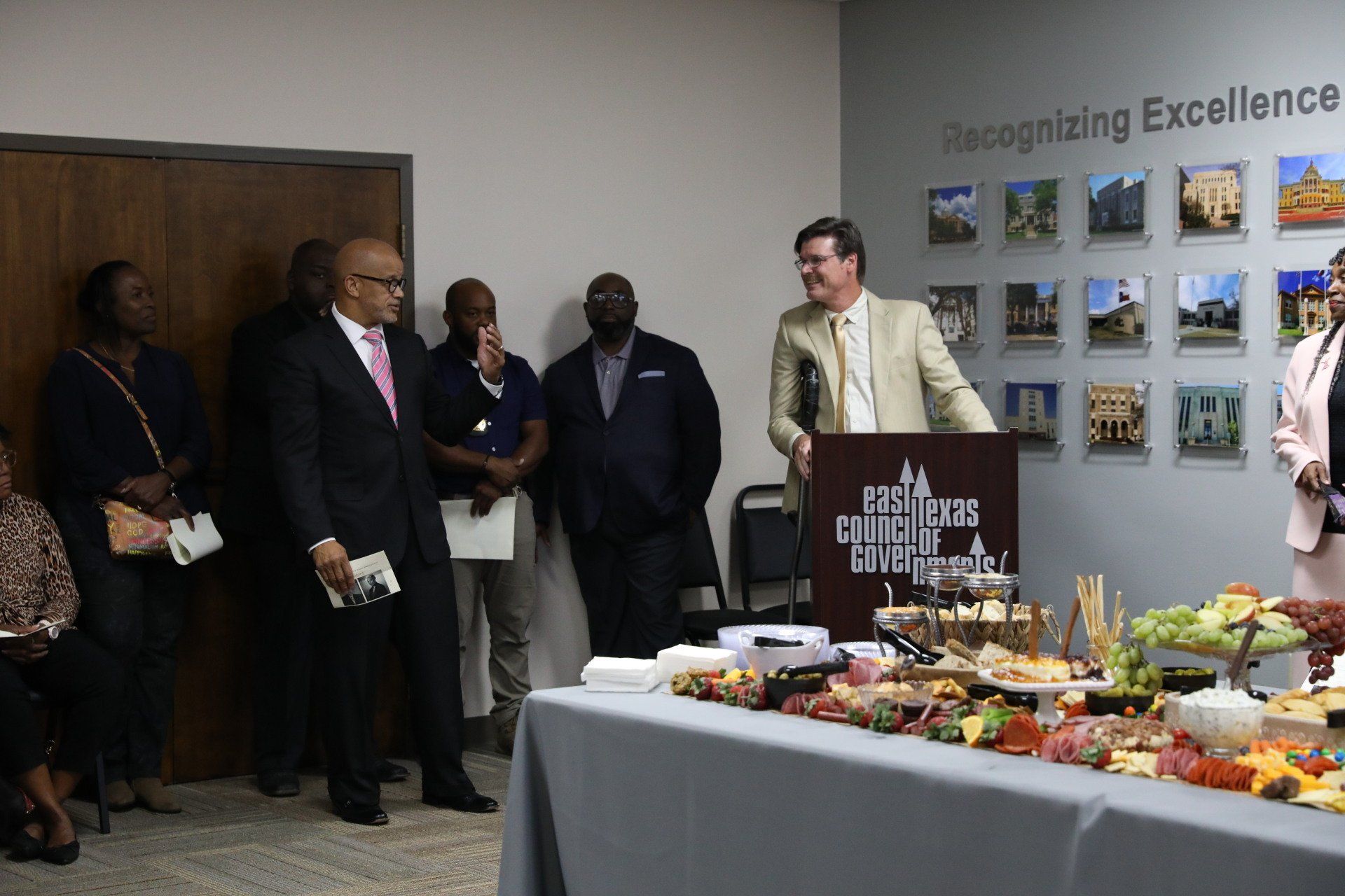 A group of people are standing around a table with food and a man is standing at a podium.