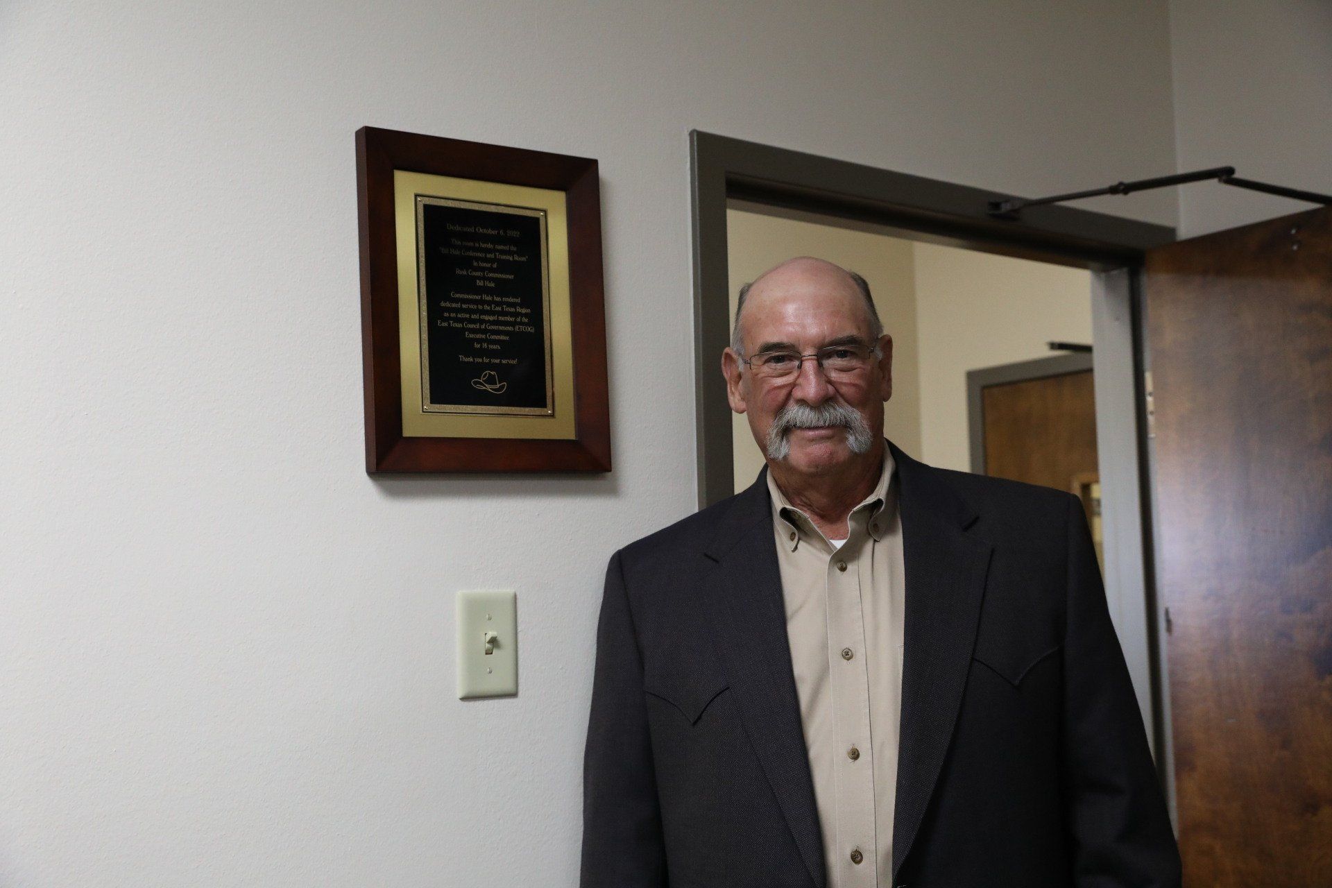 A man in a suit is standing in a doorway in front of a plaque on the wall.