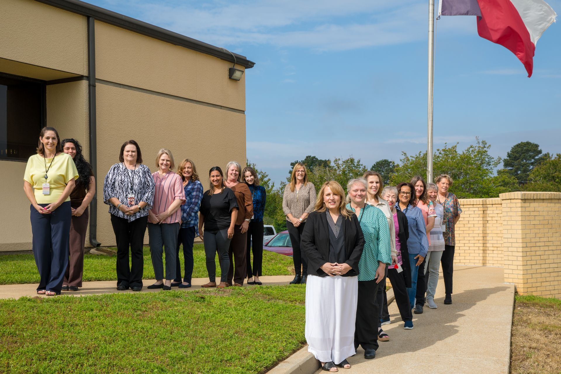 Group of people standing outside a beige building near a flagpole on a sunny day