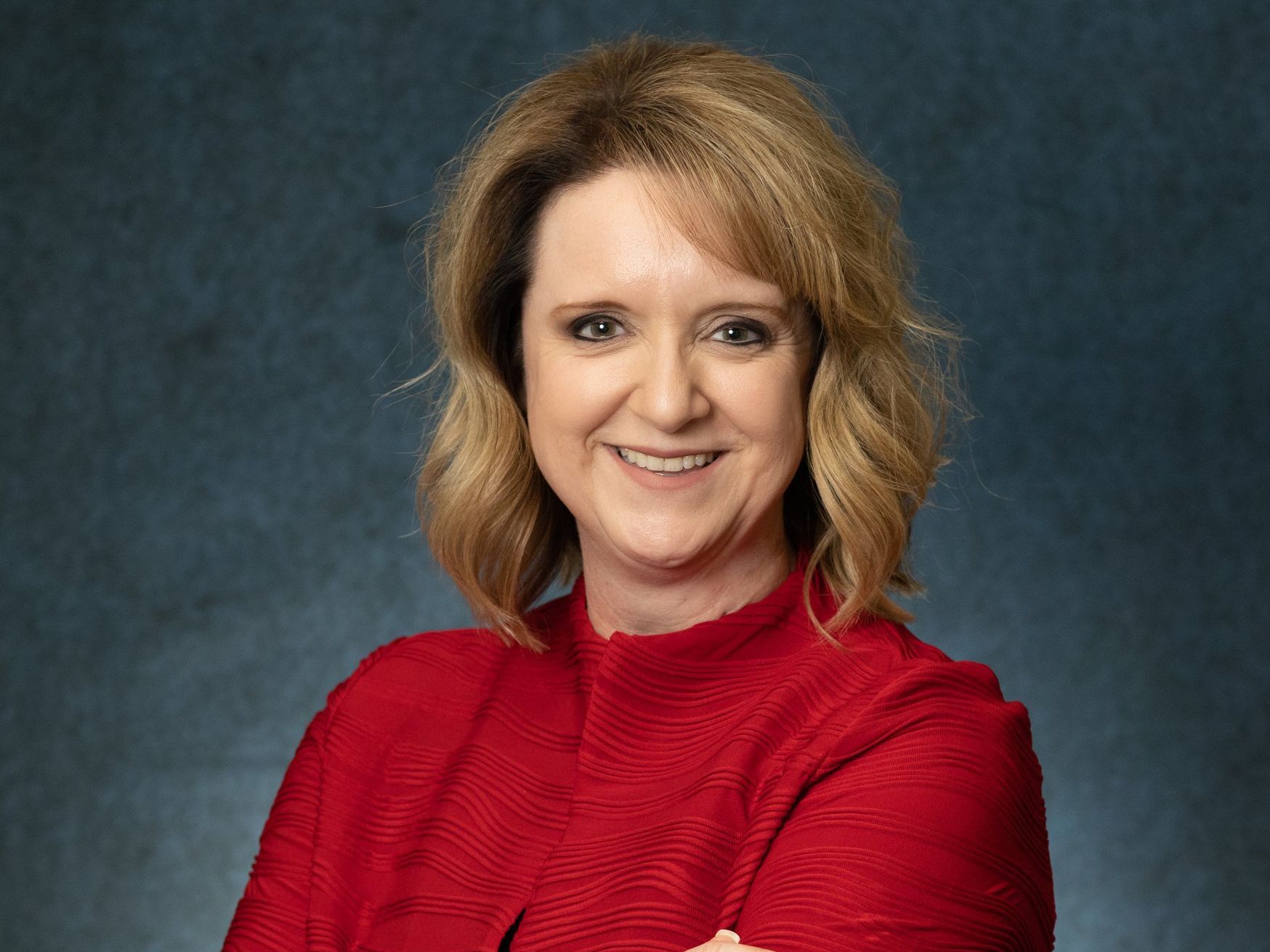 A woman in a red shirt is smiling with her arms crossed.