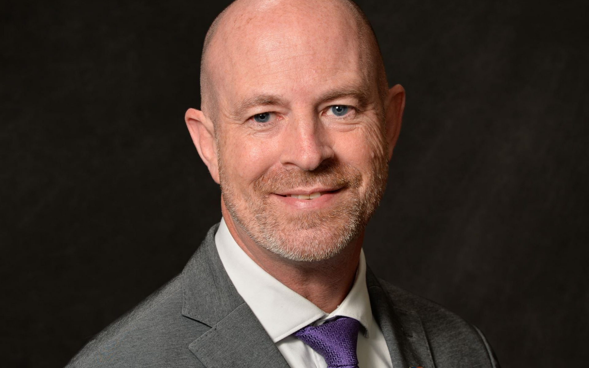 Man with fair skin and stubble in a gray suit and purple tie, smiling. Dark background.