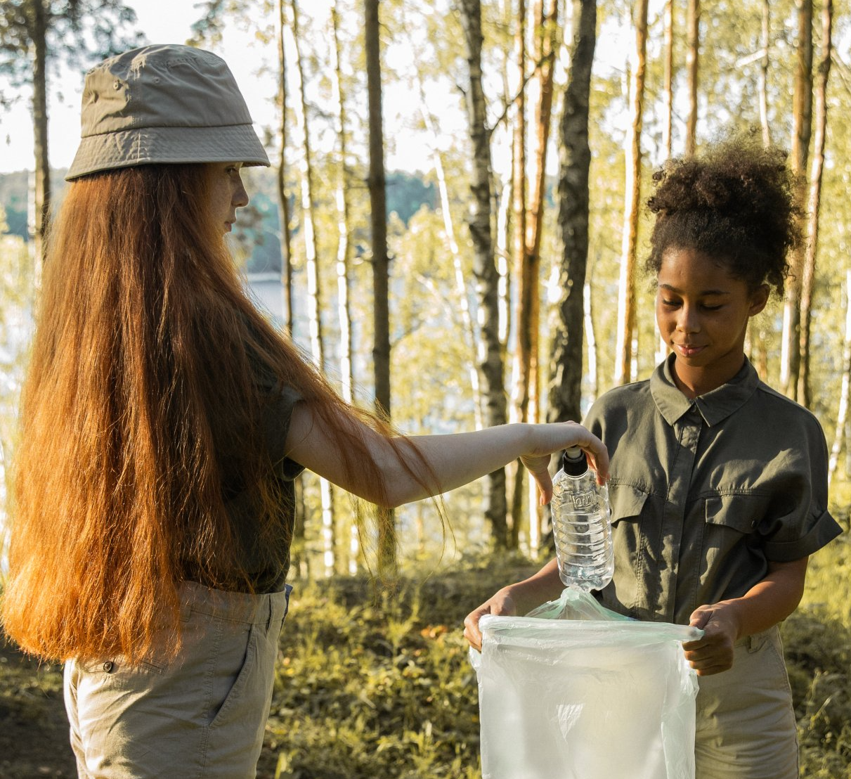 A girl is giving a bottle of water to another girl in the woods.